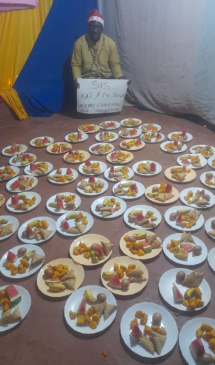 Rows of plates of samosas, watermelon, bananas, boiled eggs and snacks laid out for a Christmas 2024 meal, with a Foundation member holding a hand-written Merry Christmas thank-you sign for the supporters from Australia.