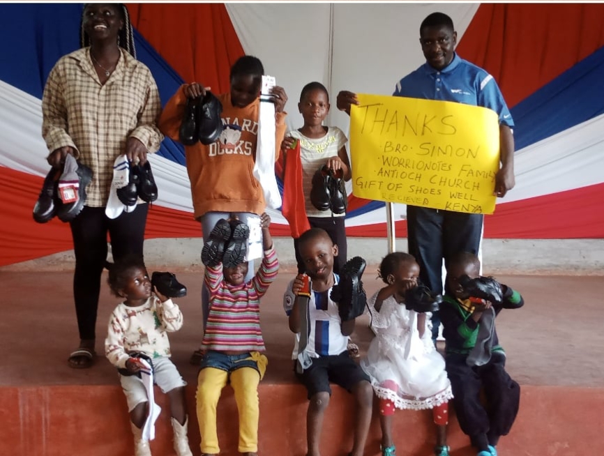 Children holding up their new school shoes and socks, with a Foundation team member holding a hand-written thank-you sign for Bro. Simon and the WarriorNotes family.