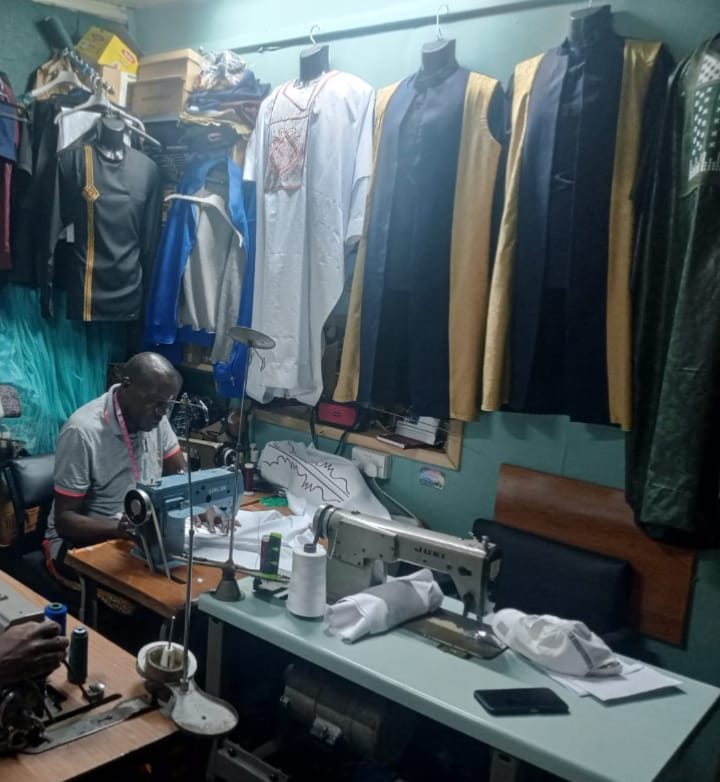 Mr Owino at work at his sewing machine in his tailoring workshop, with finished robes and shirts on display behind him.