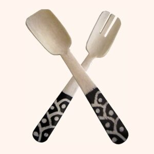 A crossed bone salad spoon and fork with black handles decorated in a white batik-style pattern.