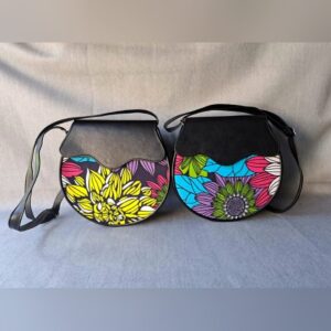 Two round crossbody bags with black leather flaps and bright floral Ankara fabric fronts, one yellow and one blue and purple.