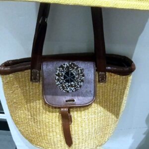 Natural sisal tote with brown leather trim and shoulder straps, and a leather flap set with a round beaded disc.