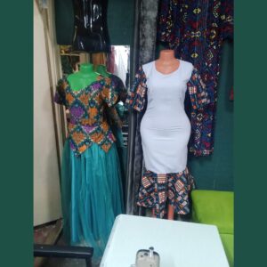 Two dresses on mannequins: one with an orange, purple and teal African print peplum bodice over a long teal tulle skirt, and a fitted pale grey dress with navy and orange African print sleeves and a flared print hem.