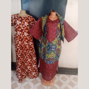 Two long African print dresses on mannequins: one in a maroon, orange and white geometric kitenge print, and one in maroon with a bold multicolour geometric print panel, wide sleeves and a matching head wrap.