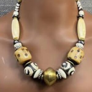A chunky necklace of cream and black batik bone beads with dotted and swirl patterns, large carved square beads and a round brass bead at the centre, shown on a display bust.