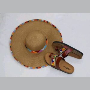 A wide-brimmed natural straw sun hat with colourful triangle beadwork around the crown and brim edge, beside a pair of brown leather toe-loop sandals with a matching beaded strap.