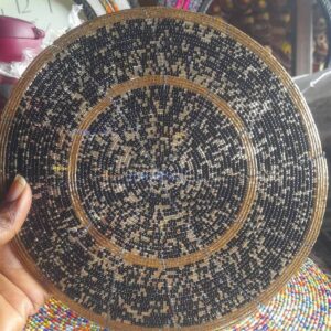 A large round beaded table mat in black and gold patterns with a gold border, held up in front of a second round mat beaded in bright multicolour.
