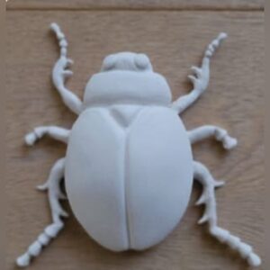 A white sculpted beetle with a smooth domed shell and finely detailed jointed legs and antennae, lying on a wooden surface.