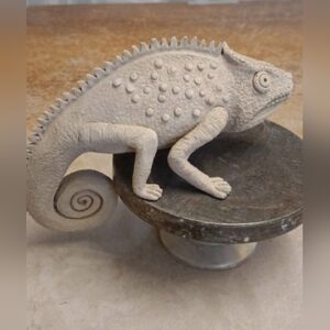 A handmade stone-grey ceramic chameleon with a crested back, dotted skin and a tightly curled tail, perched on a dark round stand.
