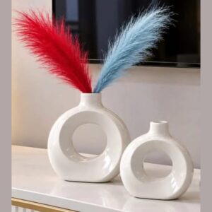 Two glossy white ceramic donut-shaped vases, one larger holding red and blue pampas grass stems, on a white marble surface.