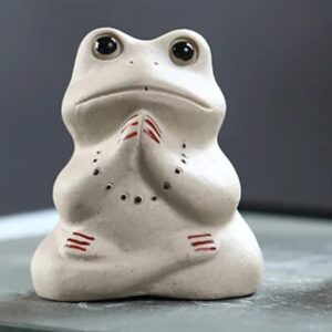 A small cream ceramic frog sitting cross-legged with its hands pressed together as if praying or meditating, with dark glossy eyes and red painted stripes on its hands and feet.