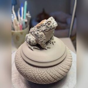 A round stoneware jar with a hand-carved crosshatch texture and a lid topped by a detailed sculpted toad.