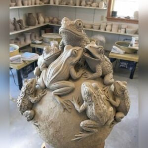 A natural clay sculpture of a round globe covered in finely detailed frogs climbing over one another, photographed in a pottery studio.
