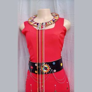 A complete outfit on a mannequin: a fitted sleeveless red dress with hanging silver chains, a wide Maasai beaded collar with long coloured bead strands, and a wide black leather belt with beaded medallions and draped silver chains.