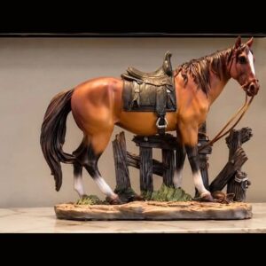 A detailed sculpture of a chestnut horse with a dark mane, a saddle and reins, standing beside a weathered wooden fence on a rocky base.