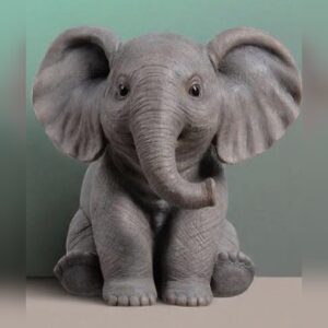 A grey statue of a baby elephant sitting on its haunches with large ears spread wide and its trunk curled upwards, with finely detailed wrinkled skin.