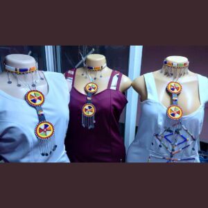 Three fitted sleeveless dresses on mannequins, in white, maroon and white with a beaded diamond pattern, each styled with a Maasai beaded choker and a long pendant of round multicolour beaded medallions with silver chain fringe.