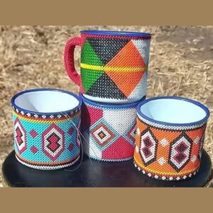 Four white enamel mugs with blue rims, each covered on the outside in colourful Maasai beadwork in geometric patterns of green, orange, turquoise, red, black and white.