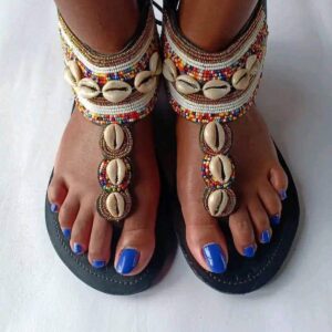 A pair of black-soled gladiator sandals worn on the feet, with wide ankle bands and toe straps covered in multicolour, white and gold Maasai beadwork and decorated with rows of cowrie shells.
