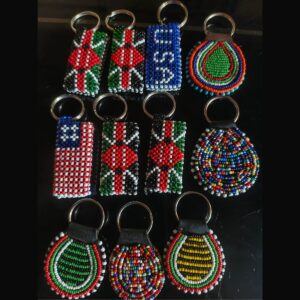 A selection of handmade beaded keychains on silver rings: rectangular designs in the Kenyan flag, the American flag and a blue pattern, and round leather-backed designs in colourful Maasai beadwork.