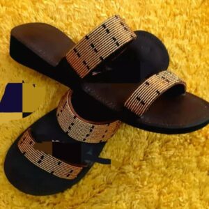 A pair of brown leather slide sandals with two wide straps covered in gold beadwork with small black accents.
