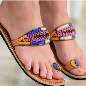 A pair of tan leather flat sandals worn on the feet, each with a wide strap beaded in pink, blue, yellow and white and a round beaded toe ring.