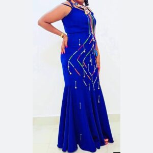 A woman wearing a long royal blue mermaid dress with thin straps, decorated with colourful Maasai beaded strands in a diamond pattern and hanging silver beaded drops, styled with a beaded collar and cuff.