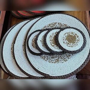 Round beaded placemats and matching coasters in pale blue-white beads with a gold beaded pattern, each edged with braided brown leather.