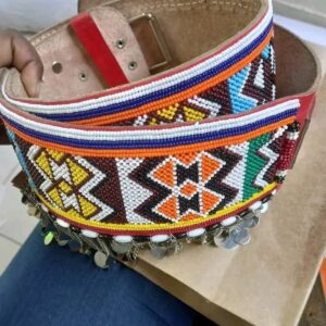 A wide tan leather belt with a buckle, covered in dense Maasai beadwork in bold geometric patterns of orange, white, brown, yellow, blue and green, with a fringe of silver chains and discs along the lower edge.