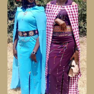 Two women in long fitted Maasai-styled dresses: one turquoise with attached cape sleeves and a black beaded leather belt with silver chains, the other purple with a wide beaded waistband, beaded strands on the skirt and a checked shuka cape.