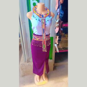 A mannequin dressed in a sleeveless lilac top and a long fitted purple skirt with a front slit, styled with a wide Maasai beaded collar with long beaded strands and a wide beaded waistband with hanging silver chains and discs.