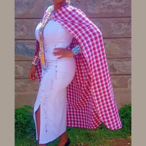 A woman wearing a long fitted white dress with a colourful Maasai beaded strip down the front and small beaded details on the skirt, styled with a pink and red checked Maasai shuka cape and beaded cuffs.