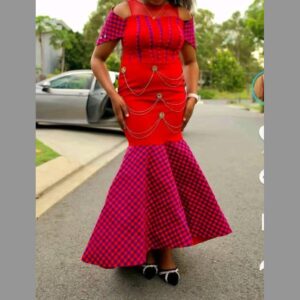 A woman wearing a long red mermaid dress with cold-shoulder checked sleeves, rows of draped gold chains across the bodice, and a flared skirt in red and purple Maasai shuka check.