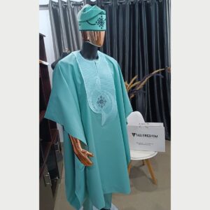 A turquoise men's agbada suit on a mannequin: a flowing outer robe over a tunic with tonal embroidery at the neck and chest, matching trousers and a matching embroidered cap.