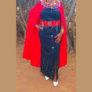 A woman wearing a long fitted navy dress decorated with small beads and hanging silver chains, a red beaded waistband with draped chains, a flowing red cape, a wide Maasai beaded collar and beaded cuffs.