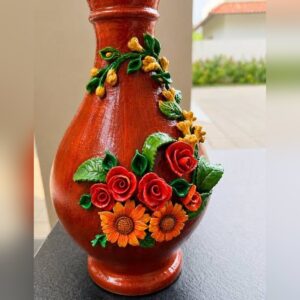 A glossy terracotta-red clay vase decorated with raised, hand-sculpted red roses, orange sunflowers, green leaves and a trailing vine of yellow buds.