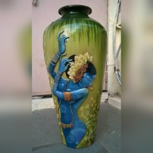A tall hand-painted terracotta vase in green with a raised figure of Radha in blue, wearing a gold flower headdress, bangles and jewellery, beside painted yellow flowers.