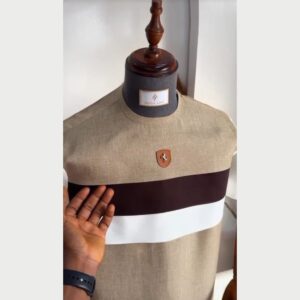 A casual kaftan on a mannequin in beige textured fabric, with bold horizontal bands of deep brown and white across the chest and a small leather badge.