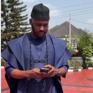 A man wearing a navy blue three-piece agbada: a wide flowing outer robe over a long tunic with sparkling embellishment down the front, with a black cap.